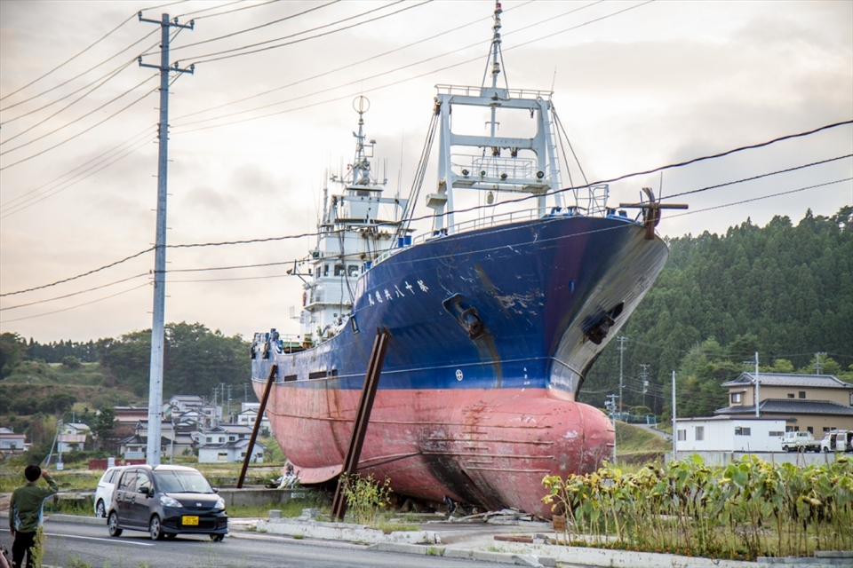 The March 2011 Pacific Coast Earthquake and Tsunami was truly devastating. I recently got to visit areas in the Miyagi province ruined by the disaster, like Kesennuma. When this ship loomed out of the dusk as we drove through the devastated city, I could do nothing but gasp and a cold feeling washed over me. Asking to stop the car, I got out and noticed the ship completely blocked off an entire side road, lying across it like an eerie blockade, surrounded by a makeshift shrine with flowers. I couldn’t grasp the sheer vastness of it but my Japanese colleague explained it had been one of many, many ships, some even bigger, that had been catapulted inland with the almighty tsunami. The rest were removed eventually, but this one remains as a memorial, a decision that has divided local residents: some feel it is a poignant reminder of all those that lost their lives, others feel it is too painful to see it everyday.