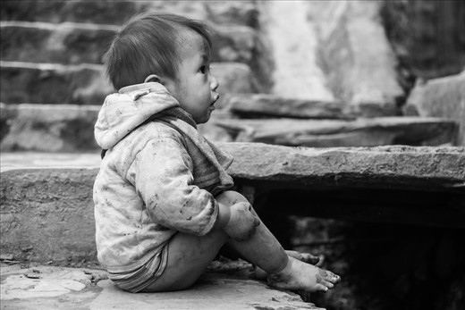 A little boy sitting on the sidewalk in front of his house looking back in.