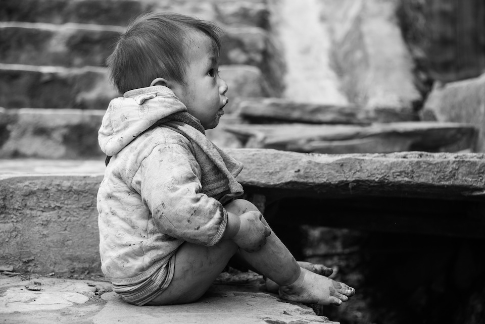 A little boy sitting on the sidewalk in front of his house looking back in.