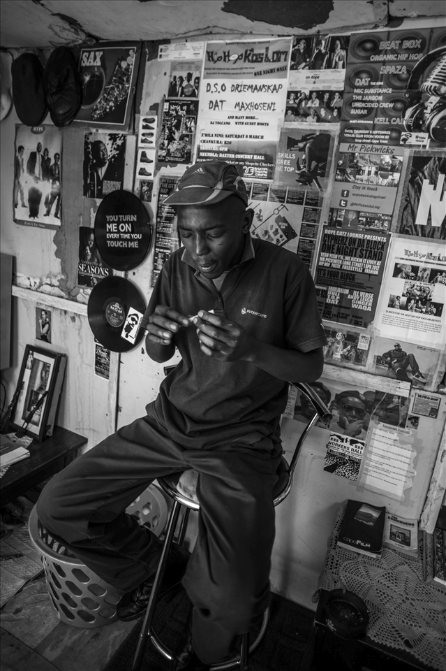 Meet Waqa. Waqa lives in Gugulethu township outside Cape Town, South Africa. After serving time in his late teens for assault, Waqa’s life now revolves around friends, smoking dope, and a passion for hip-hop music known as Spaza, a form of Xhosa township hip-hop he helped to create. Here, he rolls a dakka (marijuana) joint in his backyard shack, his love of Spaza plastered to the walls behind him.