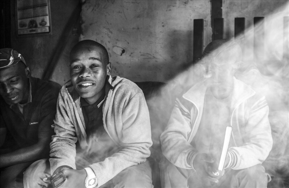 Dakka and Spaza are often found together in the township. Waqa’s friend Lucky (center) runs an outreach group that teaches both locals and tourists about traditional African music—but he also sells marijuana on the side. A shaft of light splits the smoky room. Half revealed and half hidden, part small-time crook and part community musician: like many, Lucky lives on the edge of the law, trying to make a living and still follow his dreams.