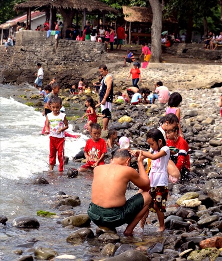 His grand daughter helping him to take a bath on the beach pouring a cup of water over his head. While other people behind are busy on their own having fun.