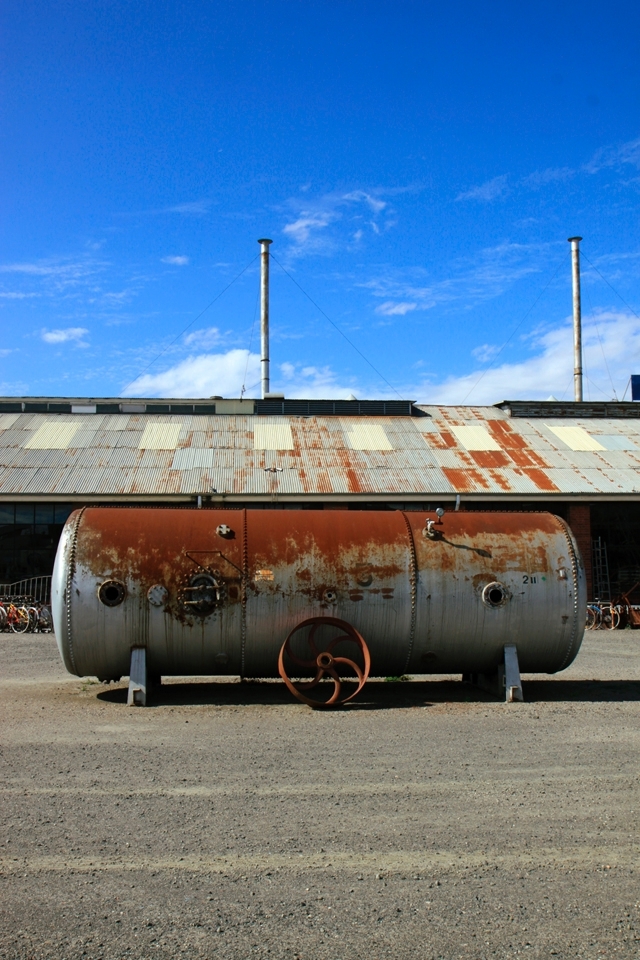 An unused tank just sitting smack in the middle of the junk yard in front of a warehouse storing various knick knacks.