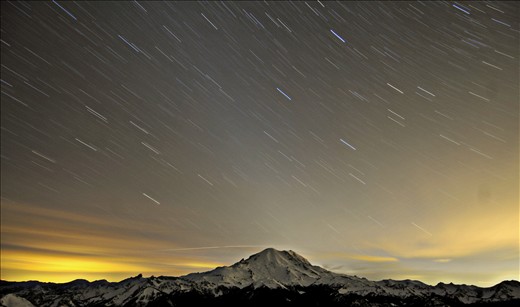 As night falls, city lights illuminate Mt. Rainier as stars tower above.