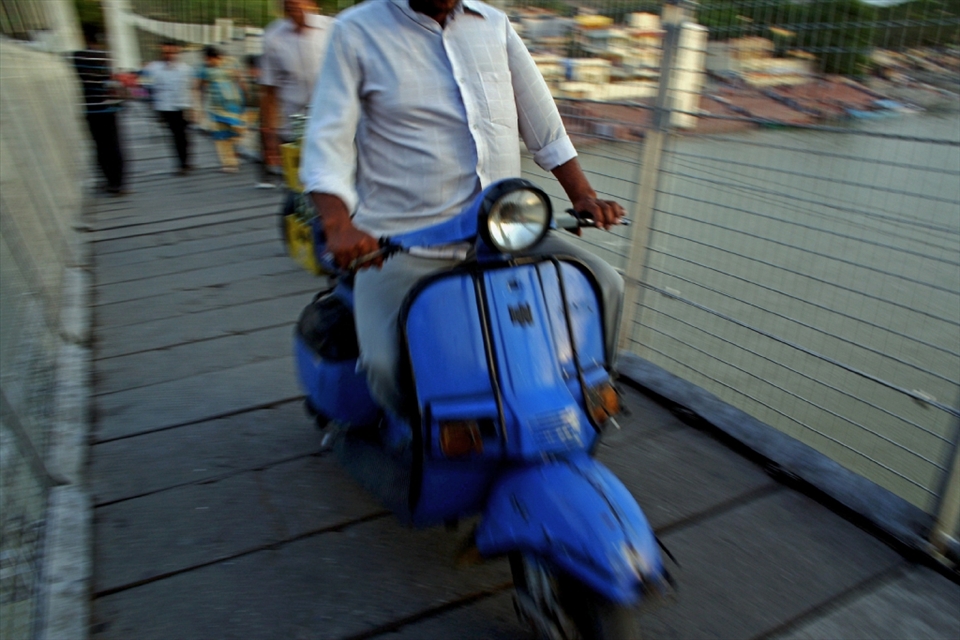 A man crosses the Laxman Jhula whilst riding a scooter, one among the more popular modes of transportation in India. The Ganges can be observed below.