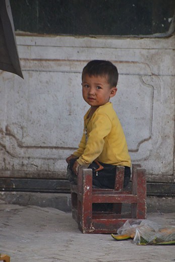 On the streets of Kashgar, a little Uyghur boy amuses himself with items he finds on the street, as his father metalsmiths a teapot over a bed of hot coal in front of his shop. 

Kashgar is an oasis city in the Taklamakan Desert of Xinjiang, and was a key post on the ancient silk road; Today there is still a lively trade of metal and clay pottery, carpets, silk, jewellery, fresh produce, and livestock.