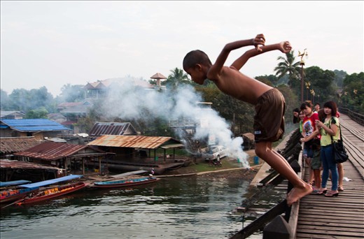 The wooden bridge in Sangkhlaburi, Thailand, is not merely a means to cross the lake by, but the centre of the community, joining 2 sides together; the Mons, and the Thais and Karens. All through the day, but particularly at sunrise and sunset, the bridge comes alive with activity; people go to and from work, vendors sell their wares, children play, monks walk to the temples, foreigners take photos. The commerce, religion, rivalry, games, tourism and natural beauty of the entire area all come together on this one wooden structure. The bridge continues to give to the community, providing an area for people to trade, socialise and play, whatever their age.  I have spent many happy hours there, sitting, watching, taking photos and soaking up the culture and diversity.

When the rainy season comes to an end, the level of water in the lake is high enough for the local children (and some foreigners if they are brave enough!) to jump off the bridge and into the water below. This group of boys were leaping off, climbing back up, discussing tactics for their next jump and showing off for my camera for at least an hour. They loved seeing the photos afterwards as well! The background shows the floating villages prevalent on the lake and also an audience!