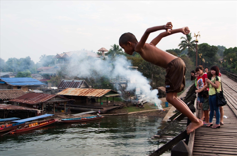 The wooden bridge in Sangkhlaburi, Thailand, is not merely a means to cross the lake by, but the centre of the community, joining 2 sides together; the Mons, and the Thais and Karens. All through the day, but particularly at sunrise and sunset, the bridge comes alive with activity; people go to and from work, vendors sell their wares, children play, monks walk to the temples, foreigners take photos. The commerce, religion, rivalry, games, tourism and natural beauty of the entire area all come together on this one wooden structure. The bridge continues to give to the community, providing an area for people to trade, socialise and play, whatever their age.  I have spent many happy hours there, sitting, watching, taking photos and soaking up the culture and diversity.

When the rainy season comes to an end, the level of water in the lake is high enough for the local children (and some foreigners if they are brave enough!) to jump off the bridge and into the water below. This group of boys were leaping off, climbing back up, discussing tactics for their next jump and showing off for my camera for at least an hour. They loved seeing the photos afterwards as well! The background shows the floating villages prevalent on the lake and also an audience on the bridge!

