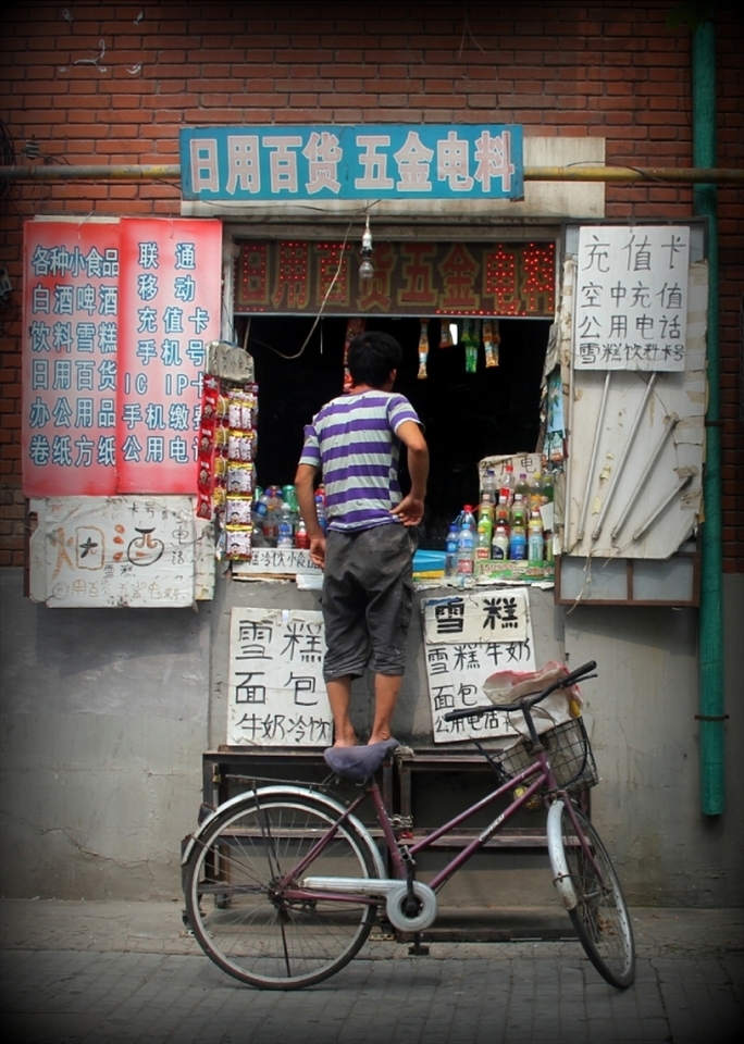 This photo really encompasses the daily life of China. With the ever present bike in the foreground and a small convenience stall operating from a window in a building in the background. These small stalls, while common enough, are starting to face to competition from big Western brand names such as Walmart and 7-Eleven. 