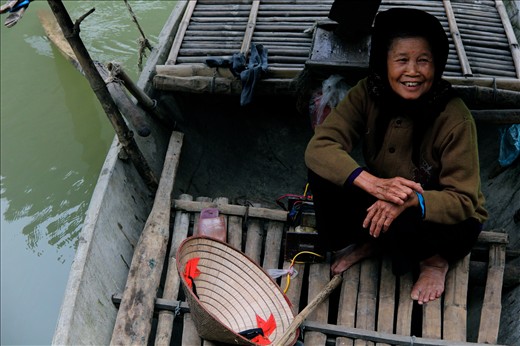 A woman sits in her boat, accompanying her 75-year-old fisherman husband. Kenh Ga is highly self-sustainable, with fish in the water, animals roaming the land, and rice fields bordering homes.