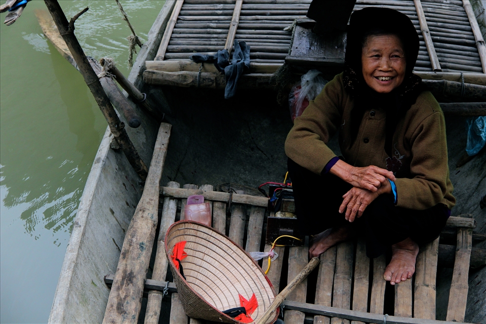 A woman sits in her boat, accompanying her 75-year-old fisherman husband. Kenh Ga is highly self-sustainable, with fish in the water, animals roaming the land, and rice fields bordering homes.