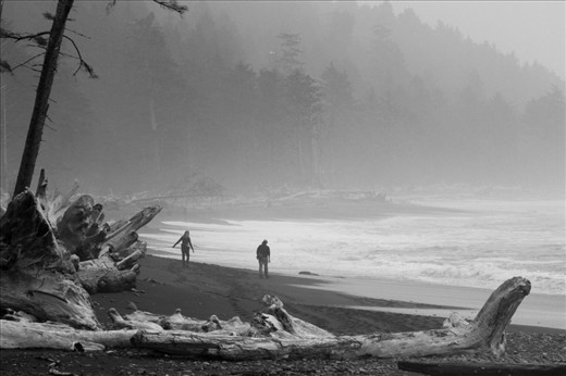 A couple walk along Rialto Beach early in the morning on a cold fall day. Fog obscures the distant land and sea.