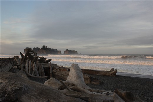 Dawn on Rialto Beach. As the sun kisses the top of the waves the mainland remains shrouded in early morning shadow. Hints of blue sky quickly dissipate as clouds roll in from offshore. A shelter is visible providing some relief from the wind and rain later that day.