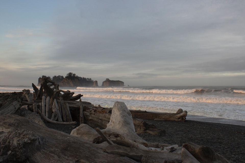 Dawn on Rialto Beach. As the sun kisses the top of the waves the mainland remains shrouded in early morning shadow. Hints of blue sky quickly dissipate as clouds roll in from offshore. A shelter is visible providing some relief from the wind and rain later that day.