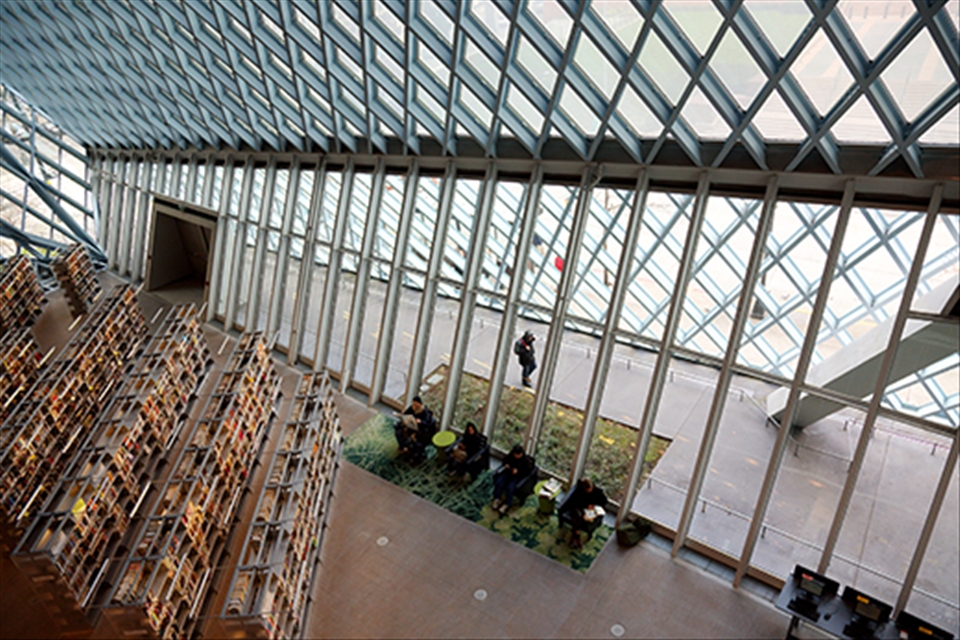A reading section is full, 4 people concentrate on reading, and a passenger walking through the hallway out side of the library. (Seattle Public Library)