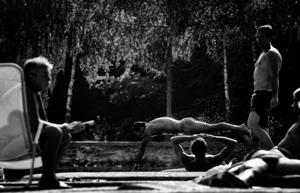 Summer: Lots of people sunbathing but only two people adventure, jumping one after another, in the lake's cold mountain water.