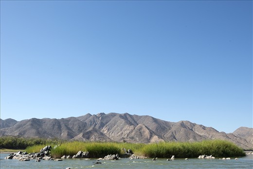 Orange River Green - In the baking summer heat, the Orange River forms a up a small strip of lush green amongst what is otherwise a desolate brown landscape. With the large catchment covering a rainfall range from 2,000mm per annum at its source to as little as 50mm per annum at its mouth, this variation creates unstable conditions which carry thousands of tonnes of sediment every summer wet season. The significant volumes of sediment makes the mouth of the river a hotspot for Diamond Divers. Richtersveld National Park, Southern Namibia