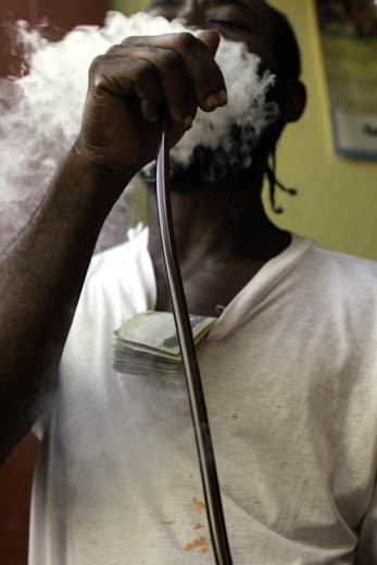 Another descendant of the Swallow Field garrison community takes a much needed smoke break from the running of his Ital Food Restaurant. He puffs on a chillum pipe with a wad of local $1000 notes purposely tucked into the neck of his t-shirt.