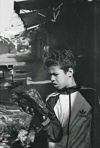 During the day of the Eid, neighborhood boys congregate in the streets of the medina (old city) to roast ram heads on bonfires built out of bed frames and wooden crates.  This street, usually crowded with goods and shoppers, becomes an apocalyptic landscape.  Smoke rises from bonfires on the unnaturally empty and boarded up street, while gangs of teenage boys compare the size of their ram’s horns and poke at the heads roasting in the fire. This boy inspects his charred ram head, perhaps imagining the taste of the brains that will show up in the couscous the next day.  