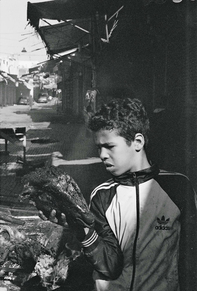 During the day of the Eid, neighborhood boys congregate in the streets of the medina (old city) to roast ram heads on bonfires built out of bed frames and wooden crates.  This street, usually crowded with goods and shoppers, becomes an apocalyptic landscape.  Smoke rises from bonfires on the unnaturally empty and boarded up street, while gangs of teenage boys compare the size of their ram’s horns and poke at the heads roasting in the fire. This boy inspects his charred ram head, perhaps imagining the taste of the brains that will show up in the couscous the next day.  