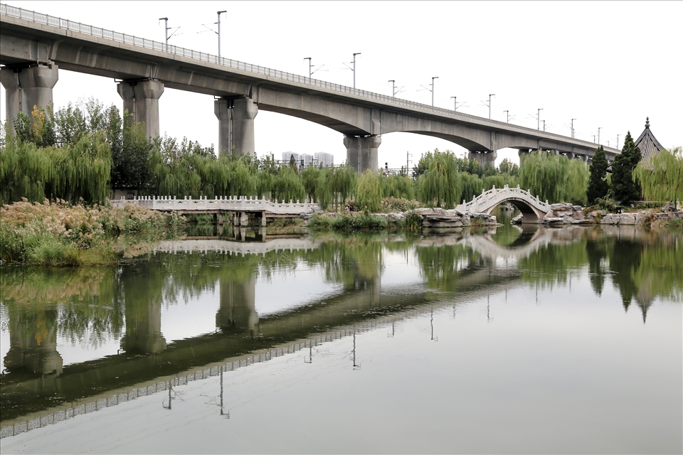 A raised railway towering over a lake and bridge on a smoggy day in a north Tianjin park. The traditional Chinese style arched bridge is dominated by the modern overbearing infrastructure. The picture reflects a lot of the conflicts China has between preserving its culture and striving for development.    