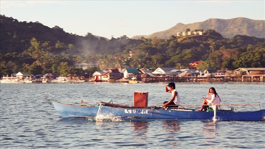 Nearing the end of a long day, locals are seen aboard their boat. They use a 
long bamboo stick to turn the rudder. A portion of the town of Coron and the 
sign on the hilltop are seen in the distance.