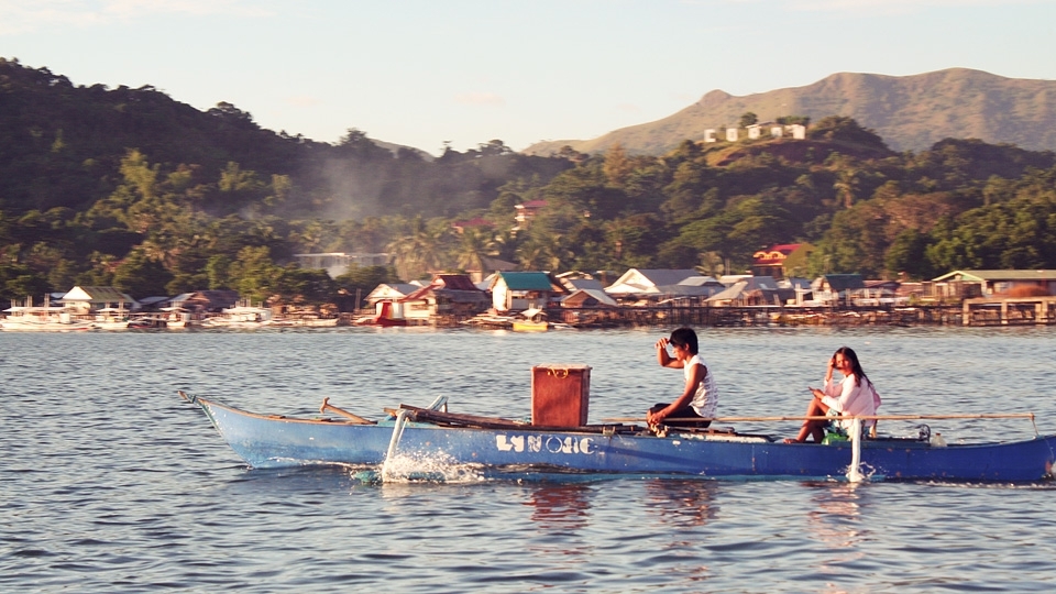 Nearing the end of a long day, locals are seen aboard their boat. They use a 
long bamboo stick to turn the rudder. A portion of the town of Coron and the 
sign on the hilltop are seen in the distance.
