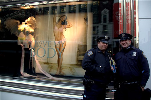 New York, United States of America, two policemen posing and holding a British boy band miniature figure in front of a provocative Victoria's Secret lingerie store window in the center of business and fashion. We live in a plastic world where looks and bank balances determine your value to the society. People are obsessed with fame and celebrities, even the police are there to entertain. Media and businesses create global trends and unrealistic expectations and try to capitalize with them in every turn, anything goes when it comes to marketing. We are slaves of market forces.