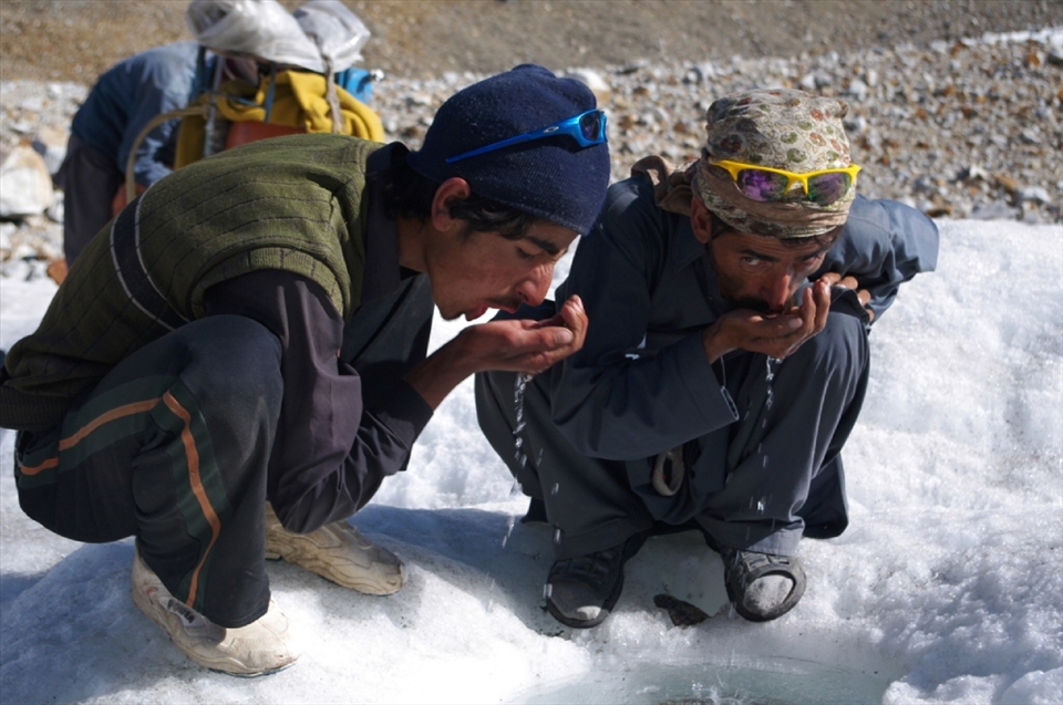 Porters wearing typical Balti attire drink from a pool on the glacier. While wages are fixed, competition to keep prices down for clients means companies frequently cut corners by not supplying adequate equipment to the men, who work in little more than cotton shalwar kameez and plastic shoes which afford them little protection from the elements.