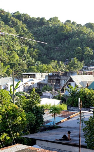 Tired of flying paper kytes, a young boy relaxes on a rooftop as the sounds of children playing and dogs barking can be heard.