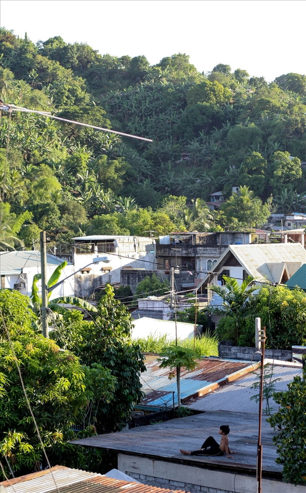 Tired of flying paper kytes, a young boy relaxes on a rooftop as the sounds of children playing and dogs barking can be heard.