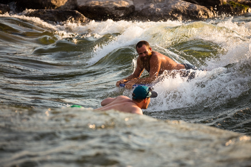 (5) Discovering new friendships from chance encounters along the Clark Fork River is commonplace among residents. People from all walks of life easily form personal connections through common interests. Here, a boogie-boarder shares a laugh with a passing “tuber” in their fleeting moments as the current sweeps them apart.  Missoula is a place where you are only a stranger but once.