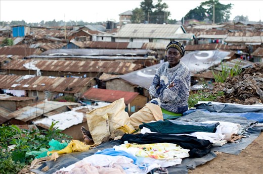 woman is selling clothes, surrounded by garbage. On backgrond Kibera village