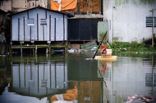 A boy uses a piece of a couch as a ship in a polluted river near an old landfill