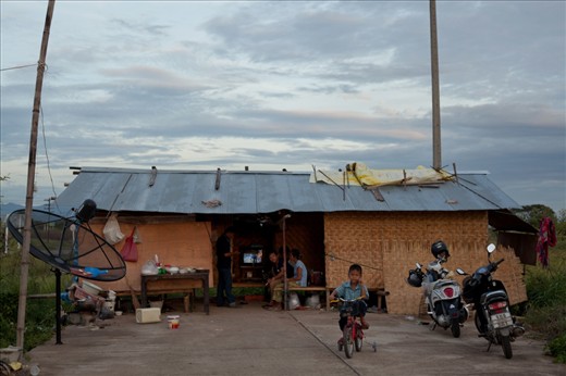 A boy plays on a bicycle outside his temporary home. Migrant workers from Shan State in Burma, this family builds makeshift housing on the construction sites in which they work. (Lamphun, Thailand)