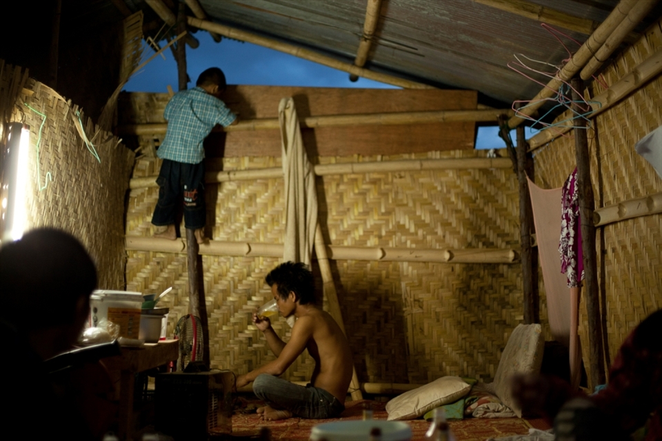 A father sips his beer as his amused son climbs the walls of the bamboo house to play with the chickens that are nesting on the roof. (Lamphun, Thailand)