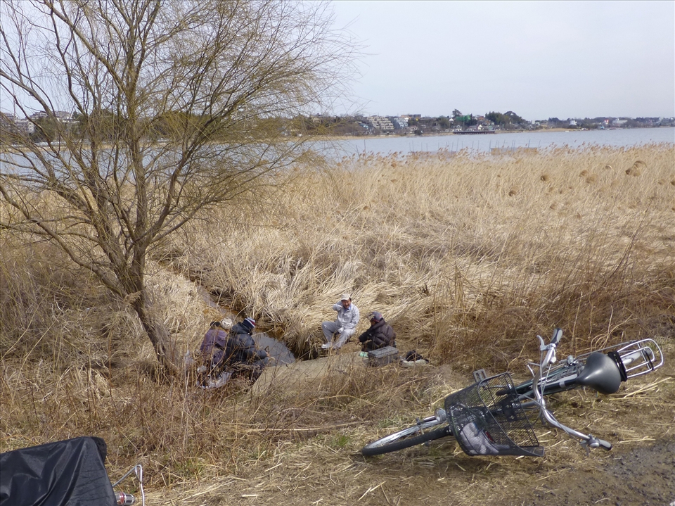 I spotted these fishermen sitting together with their rods at this little stream exiting the lake. As Caucasian foreigners such as my brother and myself are still rather unusual in Japan, particularly in smaller neighbourhood areas such as these, one of them looked up at us, chuckled and pointed us out to his friends. One can only guess what his remark was, but it could have been along the lines of, “Eh? Gaigen!” (the Japanese word for foreigner).  
Their bicycles were all casually left on the side of the road, clearly showing their means of travel and the locality of their homes.