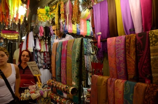 A shop owner hidden beneath swathes of hanging fabrics in bright colours in the Old City of Jerusalem. Beautiful fabrics in silk and cashmere hang outside to the attention of passing trade while 'traditional' costume hangs for the perfect tourist memento.