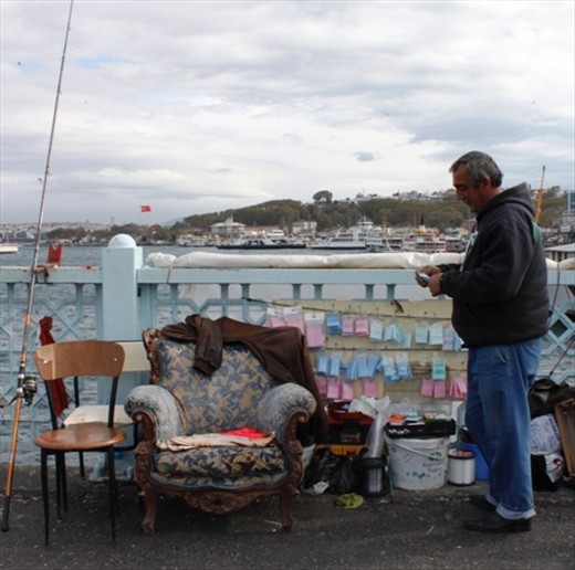 A lone fisherman is at home fishing on the Galata Bridge in Istanbul. Accompanied with a plush antique chair where he has laid his jacket, the fisherman baites his hook. During the autumnal period dozens if not hundreds gather on the bridge over the Bosphorus river as migratory fish head for warmer waters in the Aegean sea.