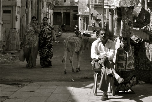 The man's sitting by his shop, and hoping for passers by to buy his work.