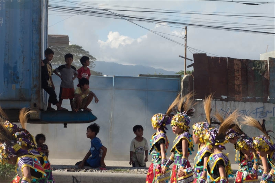 Children take cover from the blazing sun under the shadow of a large container truck as the parade begins.