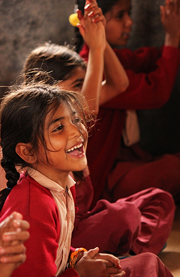 Several girls at the village's only school laugh and clap as a guest speaker tells them stories and sings with them. The girls pictured range from elementary school to high school ages as all of the village's students are taught together in a single-room building by one teacher. Each age group sits on the floor in a different corner of the school room for their studies.