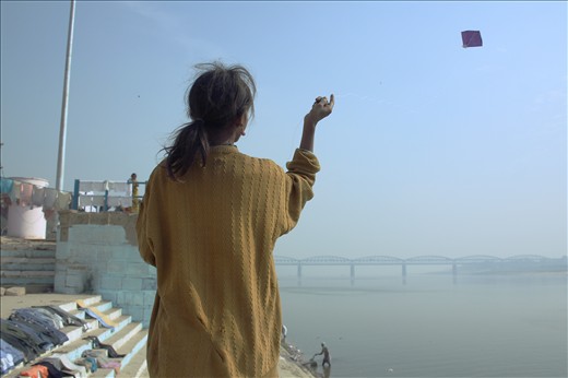 A young girl in mended jumper flies her kite peacefully over the Ganges