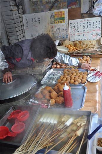 Respite in the Street.  From early morning to late in the evening, this grandma reaches over to serve, skewers fish cakes, rolls mochi, washes cups, keeps the boilers and fryers fired up and filled with comfort food cooking, all in about 60square feet of cosmic rhythm with passersby combating the cold weather. (Gimhae, South Korea)