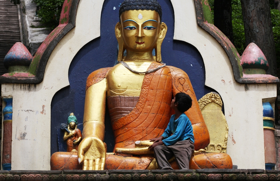 A small boy rests in the lap of a large Buddha statue at the base of Swayambhunath. 