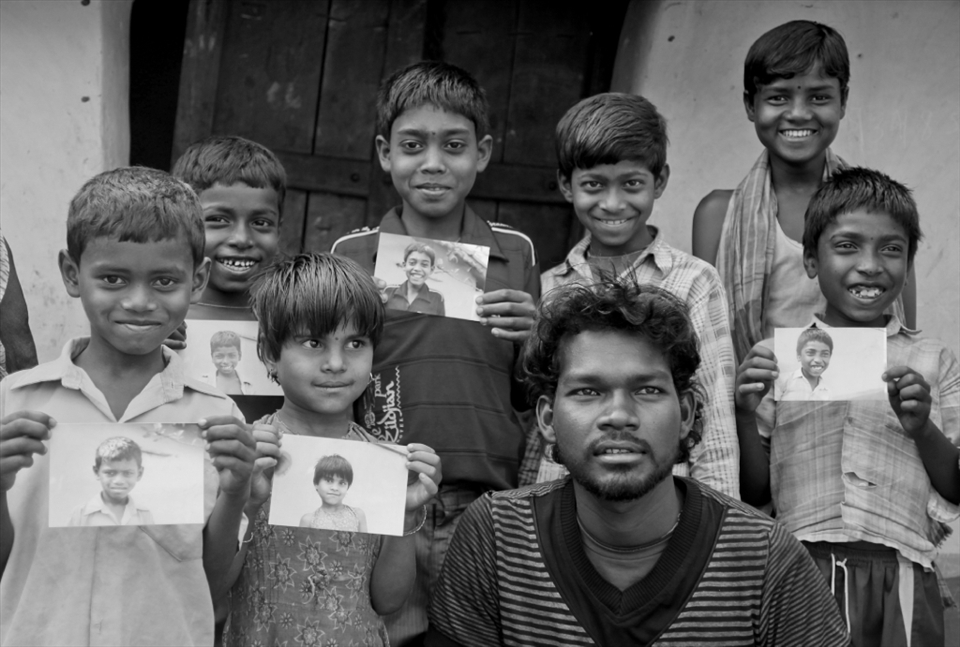 A bunch of kids along with an elder guy is all smiles, as they pose with their photographs. I had done a small Help-Portrait session with these kids on my visit to Huchukpara, whereby I gave them a print of their portraits. None of them ever had a printed photograph of themselves before and this simple gesture lit up their priceless smiles!  Notice Sujoy’s cheeky smile as he flashes his trademark unbuttoned pose in the photo.