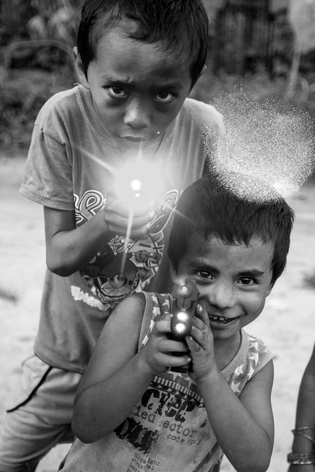 Two young boys are playing with toy guns on the street. They have been born in the Beldangi refugee camp, where they also attend school, while their families hope to migrate to a distant country.