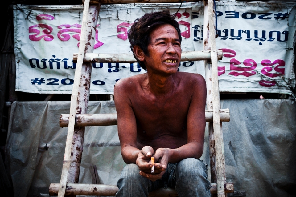 -Master of All I Survey-
In Cambodia we walked through the slums that were nearby the school we were helping at. I had a chat to this guy and asked to take his picture. He lives in the slums and tries to get work day-by-day at construction sites in order to make whatever money he could. Pictured behind him is a pretty standard “wall” of a slum house. It isn’t solid; it’s just an old advertising banner.