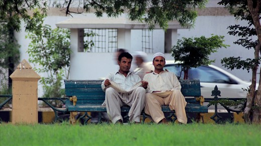 These two had just sat down on the bench near Frere Hall, Karachi, and they saw me with this really big camera, the man on the left could not resist waving at me and asking me to take a picture, which I quite eagerly did take. 
I walked up to them and showed the picture to them, they were quite happy with what they saw. Then the guy on the left, introduced himself to me, he works in one of the leather factories in Korangi. The other guy was really reserved as he did not talk much. 
The fact that they spoke to me in English was quite impressive, why they spoke to me in English was because they accidentally took me as a foreigner. The fact they spoke understandable English was again, not expected, but impressive.

What I captured was not just two people sitting in the park... I captured the common hardworking man in Karachi, trying to make a living, an honest one, quite welcoming and courteous.

After taking the picture, there was a string of violent protests in the city, I thought of these two, and how their, and the lives of many others, may have been affected by the volatile situation in the city. The two people walking behind them them made it look like their souls have escaped their bodies, quite what I think happened during the protest