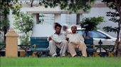 These two had just sat down on the bench near Frere Hall, Karachi, and they saw me with this really big camera, the man on the left could not resist waving at me and asking me to take a picture, which I quite eagerly did take. 
I walked up to them and showed the picture to them, they were quite happy with what they saw. Then the guy on the left, introduced himself to me, he works in one of the leather factories in Korangi. The other guy was really reserved as he did not talk much. 
The fact that they spoke to me in English was quite impressive, why they spoke to me in English was because they accidentally took me as a foreigner. The fact they spoke understandable English was again, not expected, but impressive.

What I captured was not just two people sitting in the park... I captured the common hardworking man in Karachi, trying to make a living, an honest one, quite welcoming and courteous.

After taking the picture, there was a string of violent protests in the city, I thought of these two, and how their, and the lives of many others, may have been affected by the volatile situation in the city. The two people walking behind them them made it look like their souls have escaped their bodies, quite what I think happened during the protest: by usmanbinomar, Views[721]